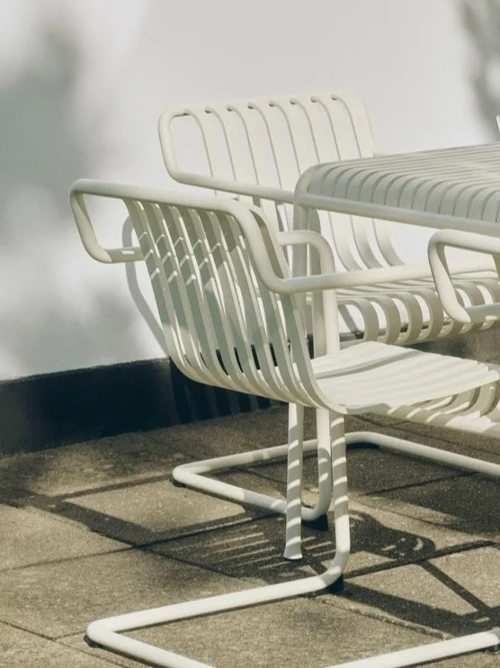 Två vita utomhusstolar i metall med ribbor och ett matchande bord på en uteplats i solljus.