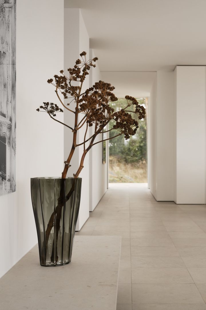 Bild som visar vasen Reed 50 cm i färgen mossgrön, formgiven av Monica Förster för Orrefors. Vasen står placerad på ett lågt sideboard och i vasen står en torkad blomma med lång stjälk och stor krona.