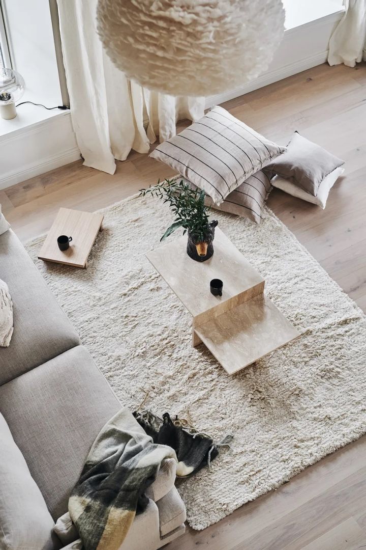 Bild som visar Cozy ullmatta från Scandi Living, en vit fluffig matta placerad i vardagsrum med grå soffa och soffbord i beige travertine.