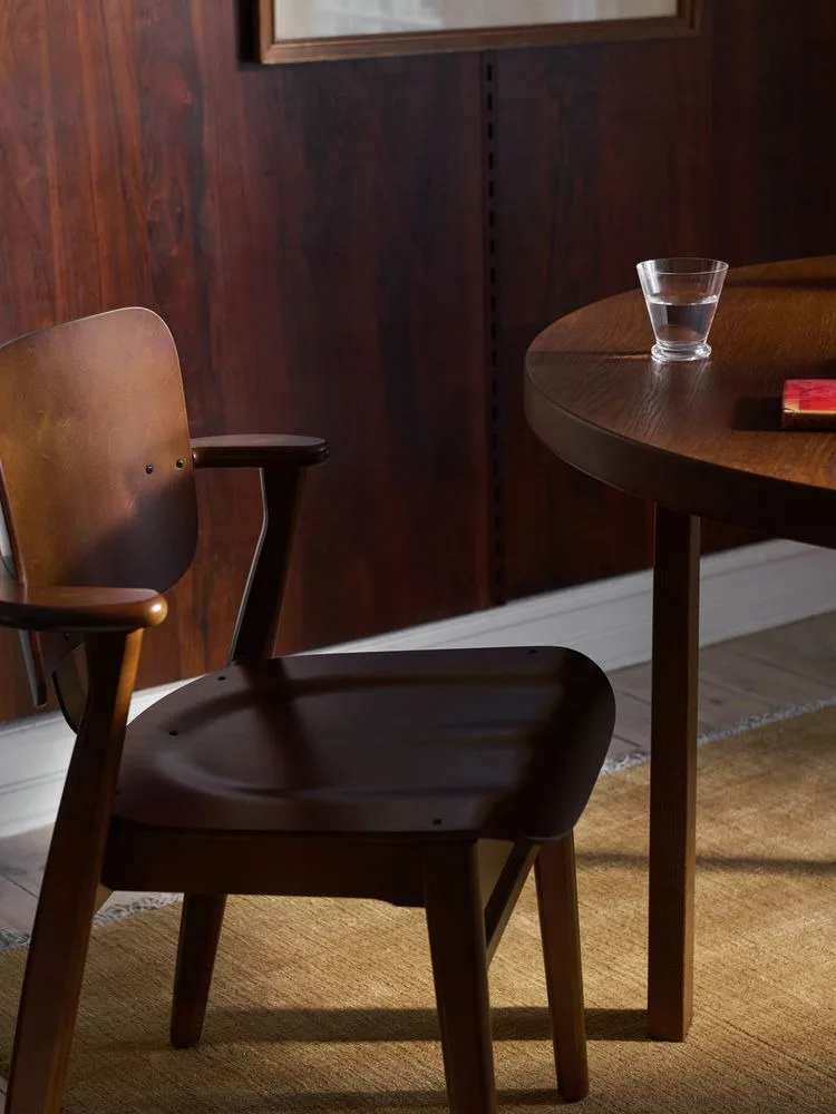 Mörk trästol med armstöd vid ett runt bord med ett glas vatten och en röd bok.