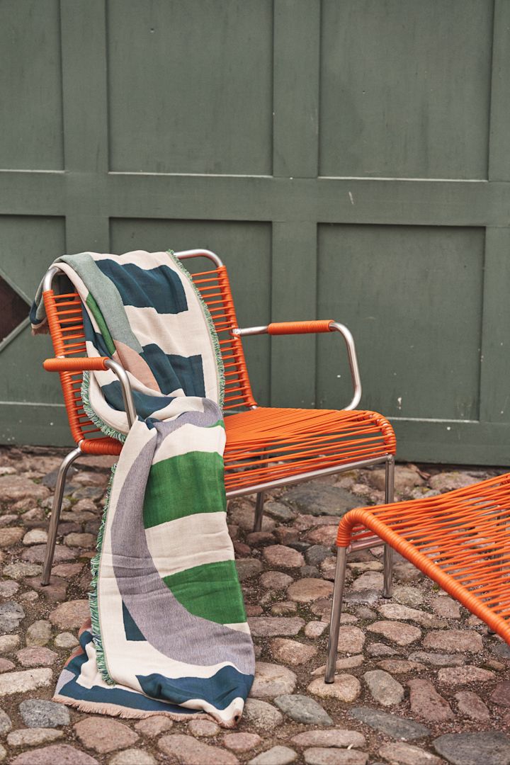 Siirto pläd är formgiven av Maija Isola för Marimekko och har ett distinkt mönster. Här hängd över orange spaghetti-stol.