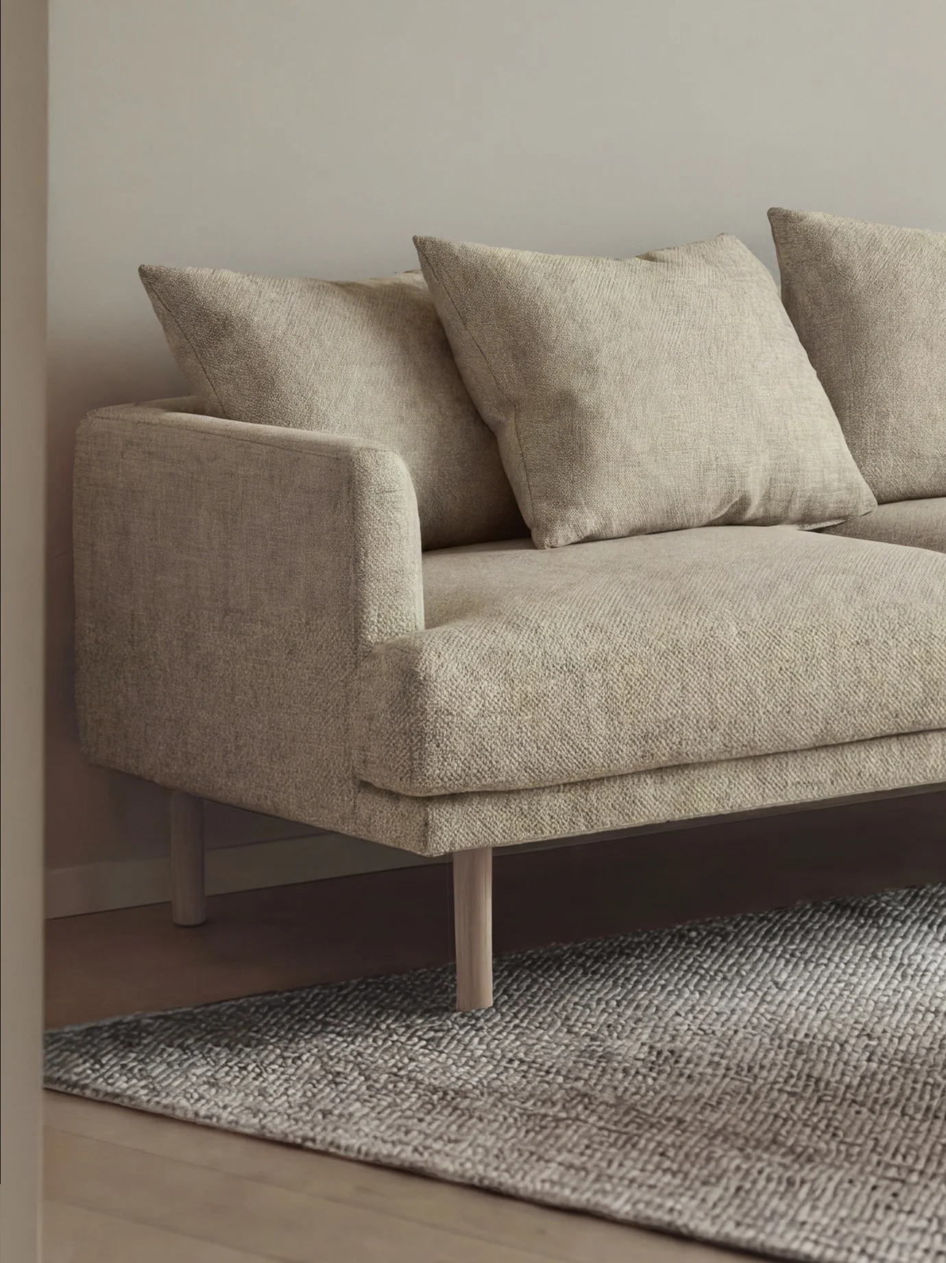 Närbild av en ljusbeige soffa med texturerat tyg, ljusa träben och kuddar, på en grå, texturerad matta.