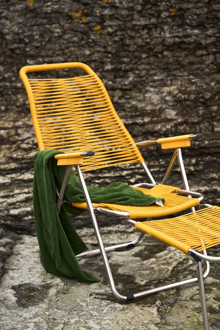 Miljöbild där Spaghetti solstol med fotstöd i gult från Fiam står på ett berg och har en grön badhandduk hängandes på ena armen.