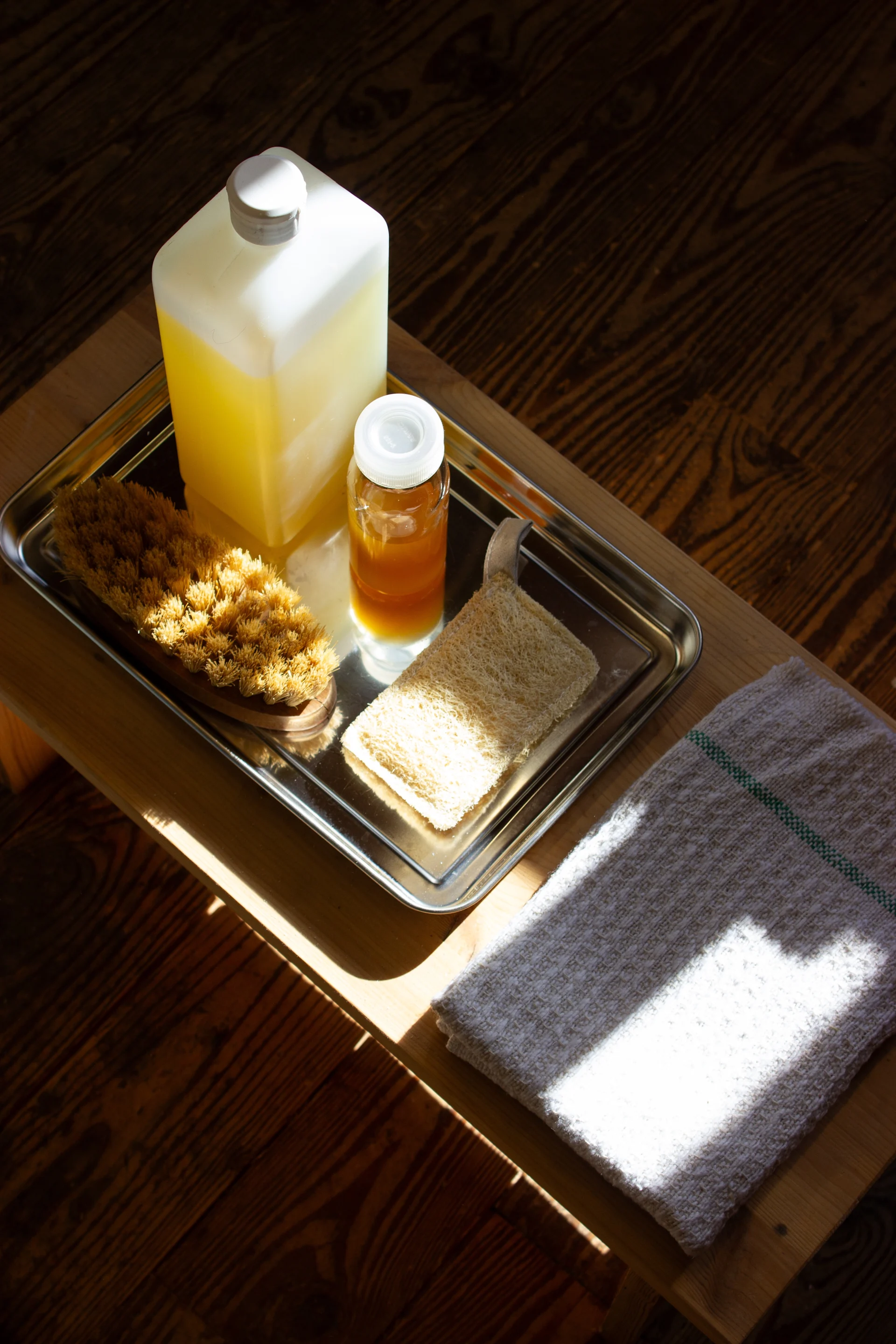 Metallbricka med gul vätskaflaska, bärnstensfärgad flaska, kroppsborste, loofah. Vit handduk, på solig träyta.