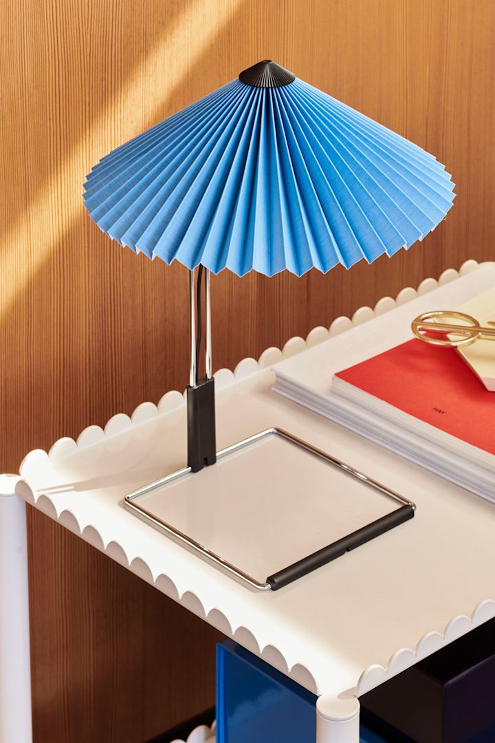 Bild som visar Matin bordslampa med plisserad skärm i blått från HAY, ståendes på vitt sidobord.