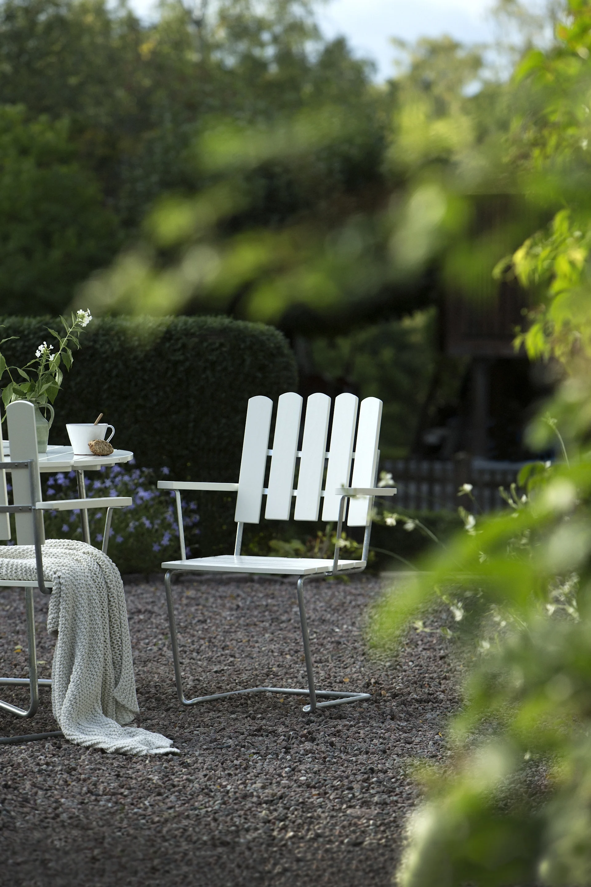 Vita ribbade utomhusstolar och bord på grus med filt och blommor i en trädgård.