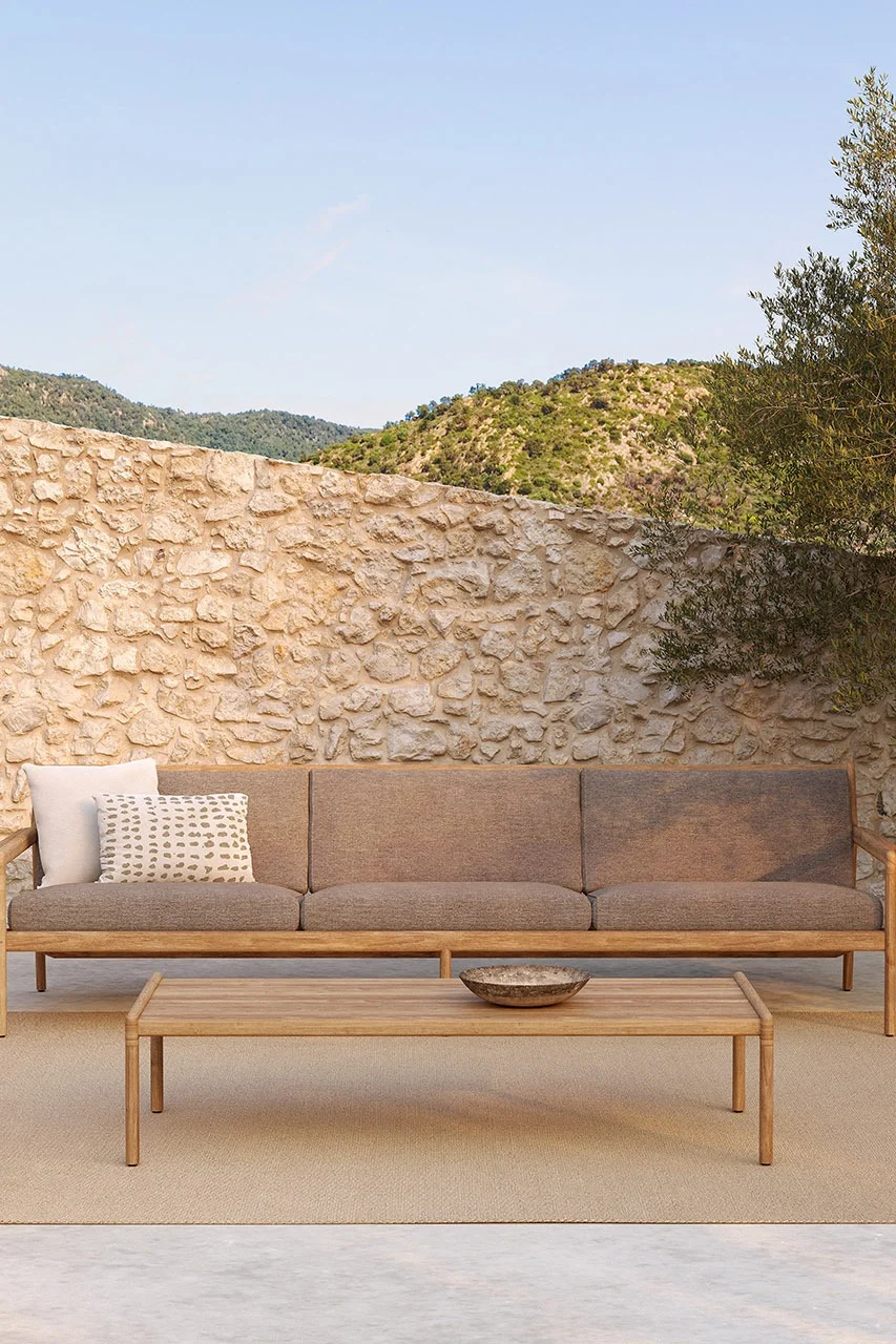 Bild som visar Jack Outdoor loungesoffa från Ethnicraft. Här med gråa dynor och stomme i teak. En stilren soffa placerad framför en hög stenmur med ett lågt avlångt bord framför, också i teak.