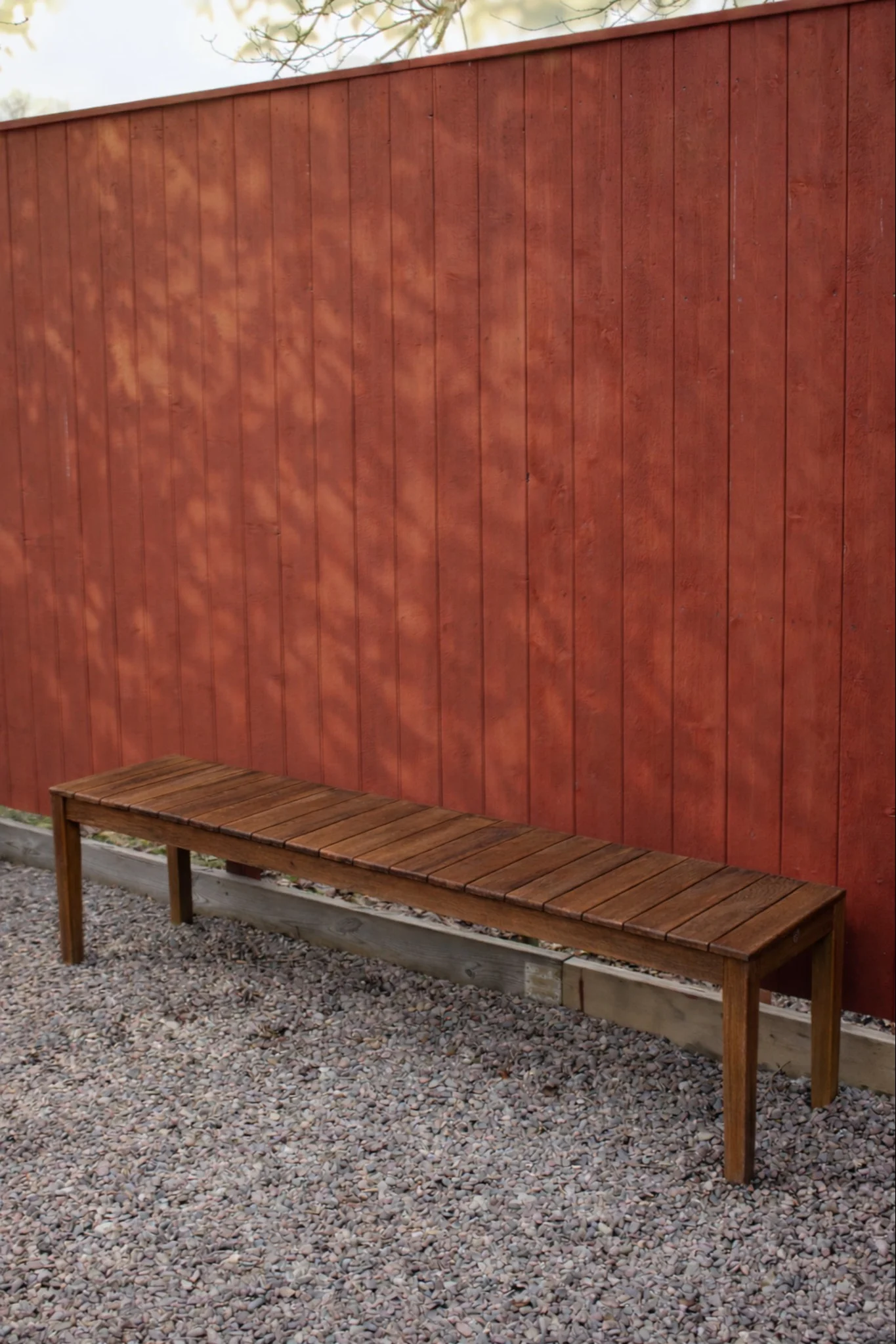 Träbänk med ribbor på grus framför ett rött trästaket med lövskuggor.