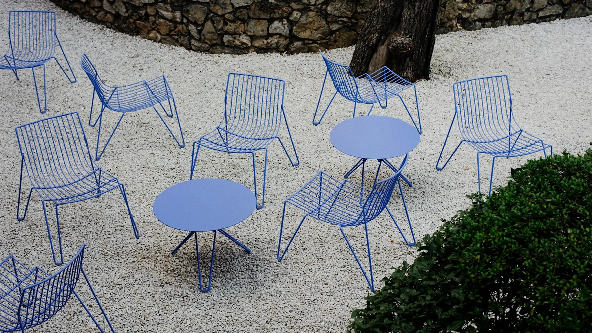 Bild som föreställer Tio easy chair loungestolar och cafébord från Massproductions, här i en blå färg där flera bord och stolar står utspridda på en grusad innegård.