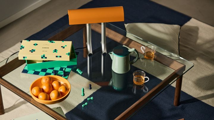 Bild som visar Anagram bordslampa i färgen charred orange stående på glasbordet Kofi, på bordet står även HAY PLAY schackspel och Sobremesa serveringsskål i gult med apelsiner i.