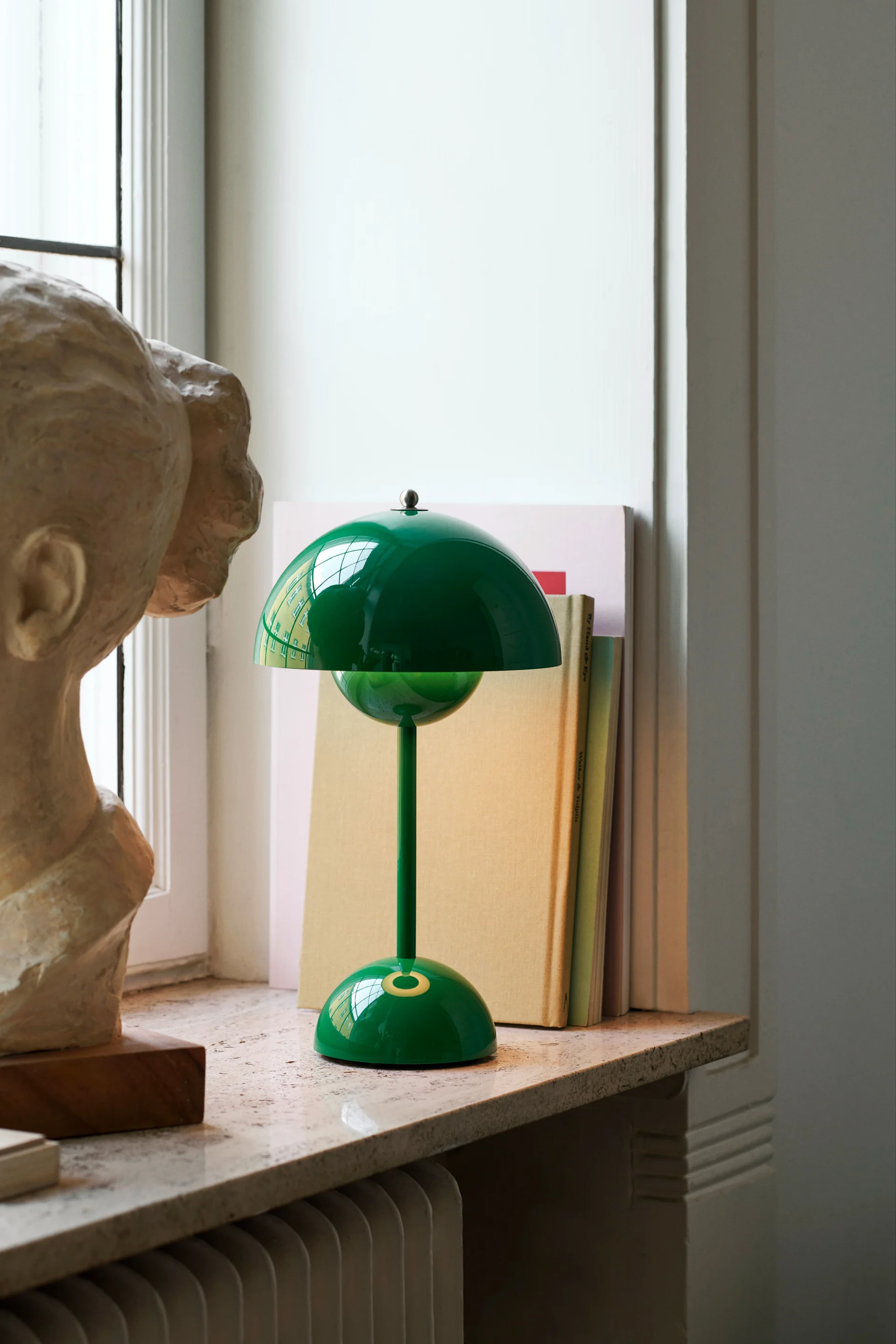FlowerPot VP9 i grönt från &Tradition är en av många populära, portabla bordslampor, här placerad i ett fönster med en dekorativ staty och böcker.