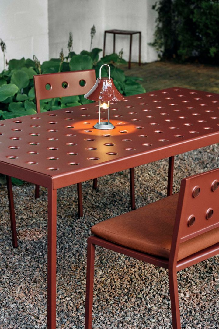 Mousqueton portabel lampa från HAY i färgen iron red, placerad på rött bord utomhus.