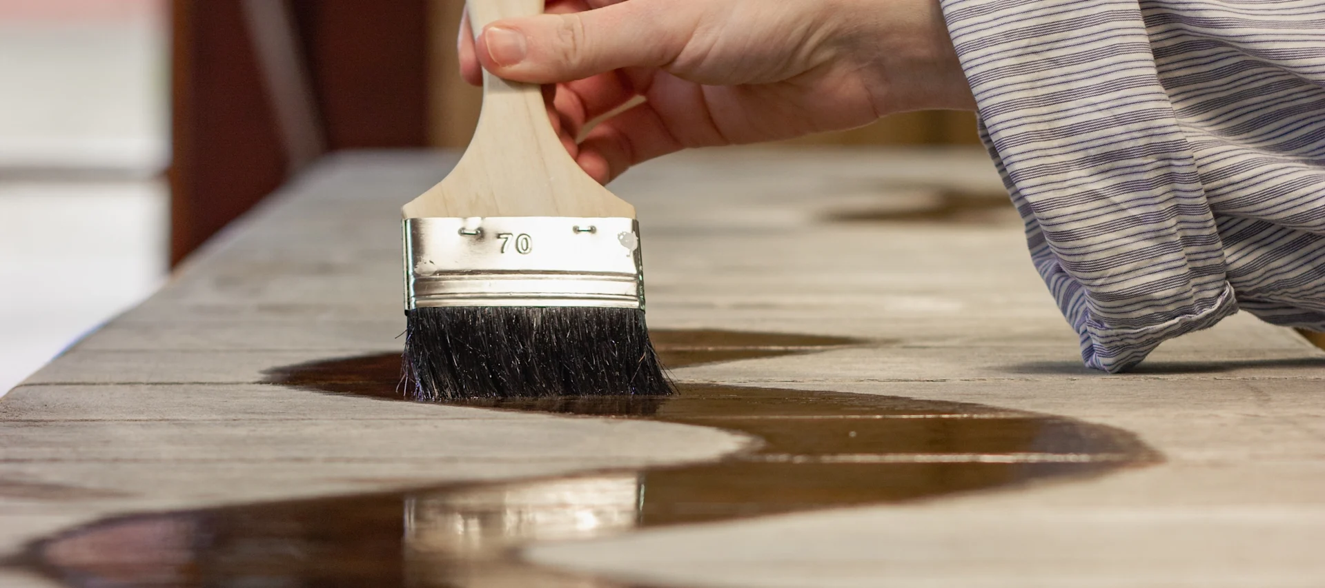 Hand med pensel applicerar vågigt, brunt betsat trä på en ljus träyta.