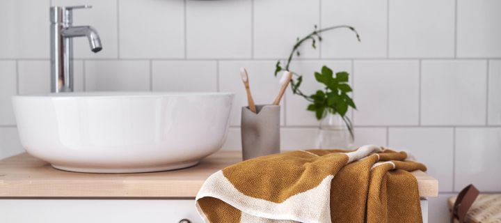 Bild som visar badrum med vitt, runt handfat, mönstrad handduk vid sidan av kommoden, en grön liten växt i vas med vit kaklad vägg i bakgrunden.