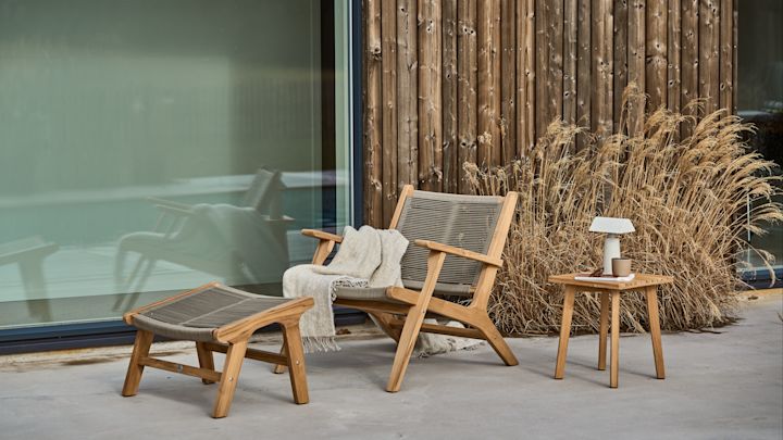 Bild som visar Sandvik loungefåtölj och tillhörande pall i teak och rep av polypropen. Möblerna står på uteplats av betong mot en mörk husvägg.