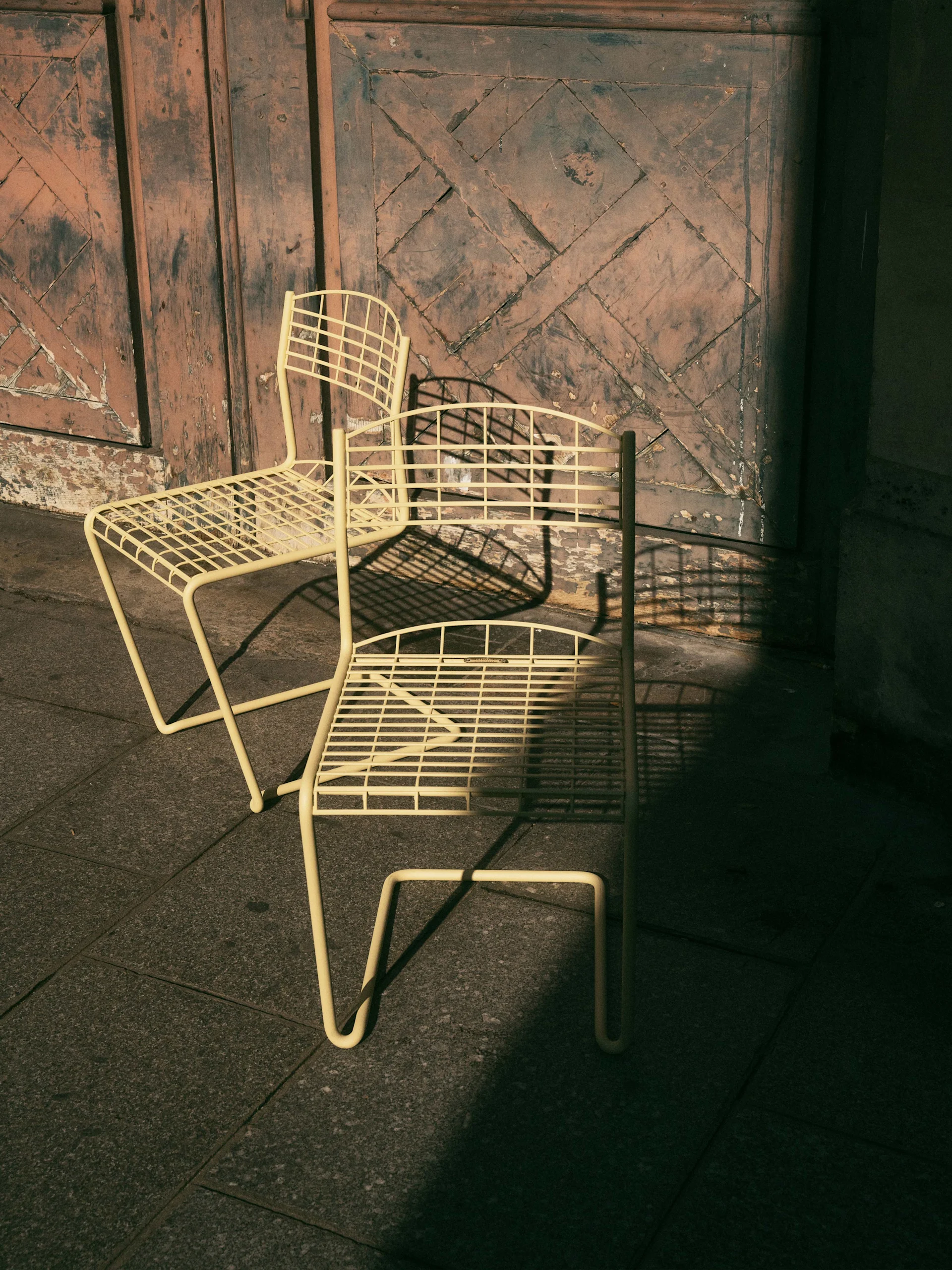 Två ljusgula metallstolar på kakel framför en sliten trädörr med geometriskt mönster, med skarpa skuggor.