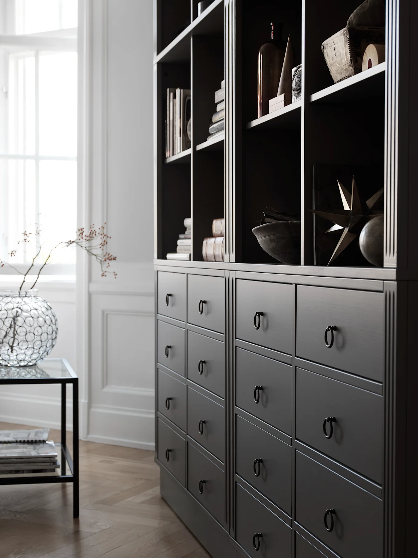 Mörkgrå bokhylla med lådor och svarta ringhandtag. Hyllorna innehåller böcker och dekorationer. Ett fönster till vänster.