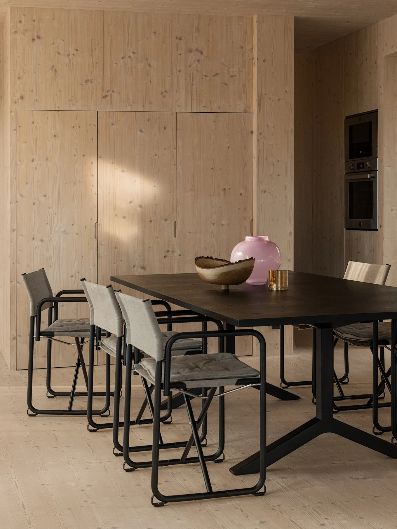 Långt mörkt matbord med åtta grå textilklädda stolar i ett rum med ljusa träväggar och -golv, med en rosa vas på bordet.