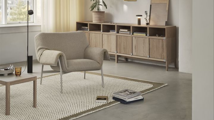 Bild som visar Wrap loungestol från Muuto placerad i ett vardagsrum. Fåtöljen är i beige med vitt stålstativ och står placerad på benvit matta med ett sidobord i trä bredvid.