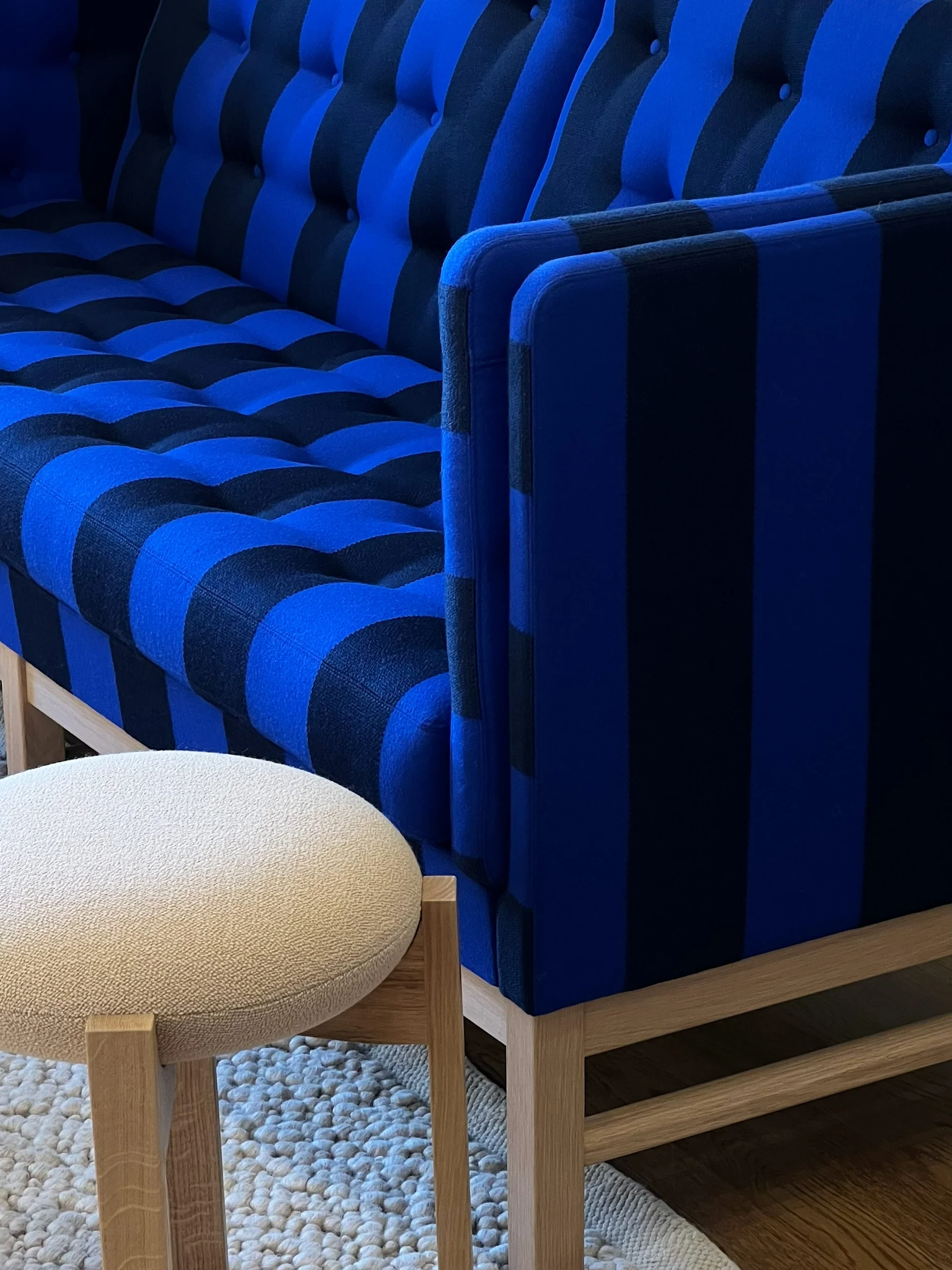 Blå och svartrandig soffa med knappar, en ljus, rund pall och en vit, texturerad matta på trägolv.