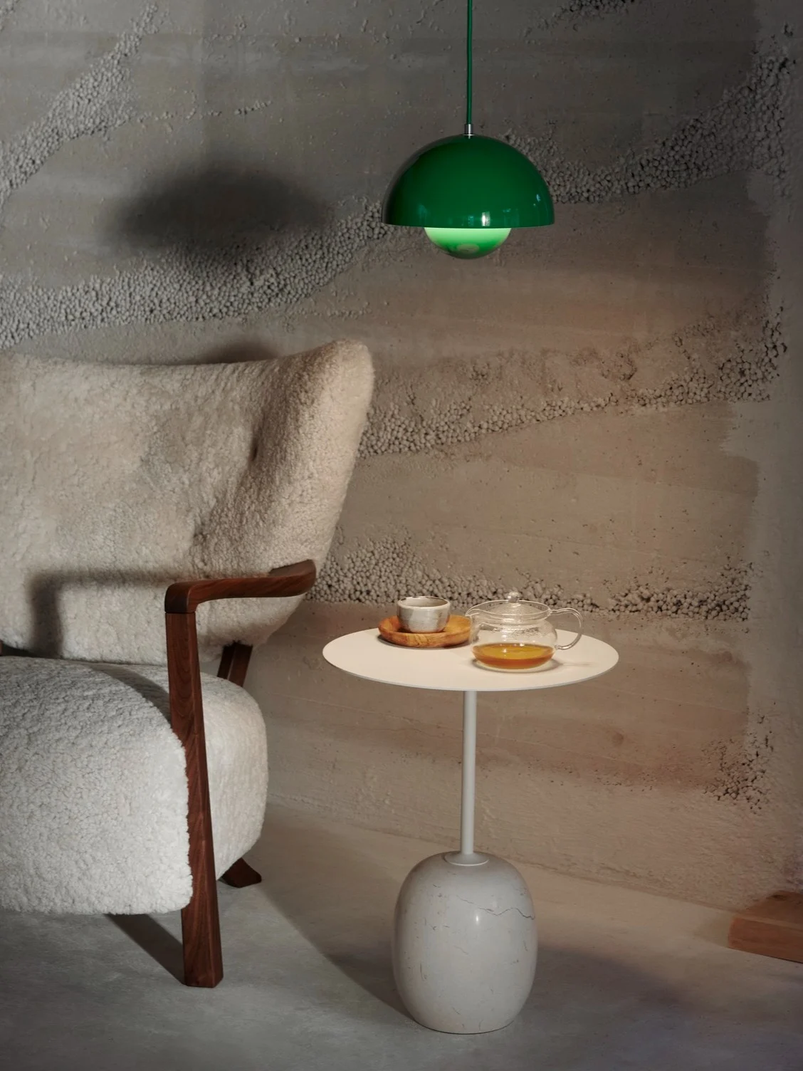 En mysig interiör med en vit, lurvig fåtölj, ett sidobord med tekanna och kopp, och en grön taklampa ovanför.