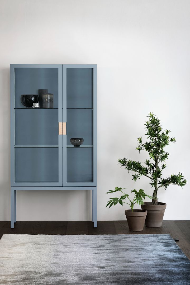 Frame vitrinskåp i färgen nordic blue från svenska Asplund, formgivet av Anya Sebton, här placerat mot vit vägg med grå matta framför och två större gröna växter i krukor bredvid på golvet.