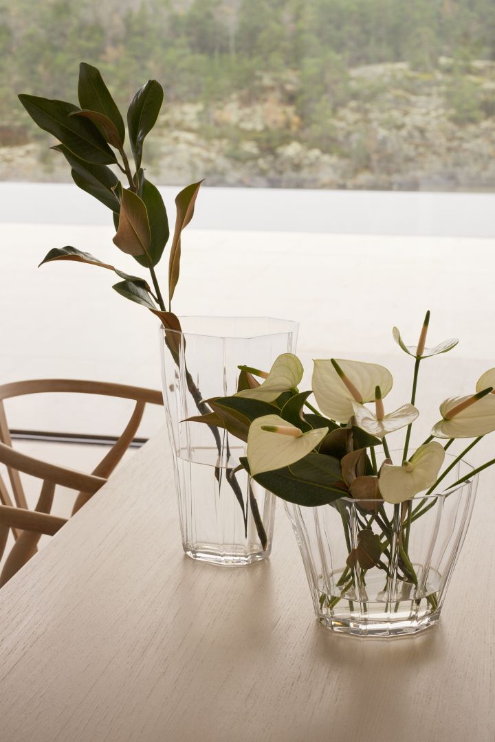 Bild som visar Reed-vaserna i storlekarna 17 cm och 30 cm i klarglas, placerade på ett köksbord i trä. I vaserna är gröna kvistar placerade. Vaserna är formgivna av Monica Förster för Orrefors.